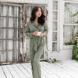 Model standing on a white wooden floor, wearing the Women's Long Sleeve Pajama Set in Gingham Fir holding a mug in front of white-paneled walls