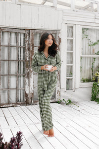 Model standing on a white wooden floor, wearing the Women's Long Sleeve Pajama Set in Gingham Fir holding a mug in front of white-paneled walls