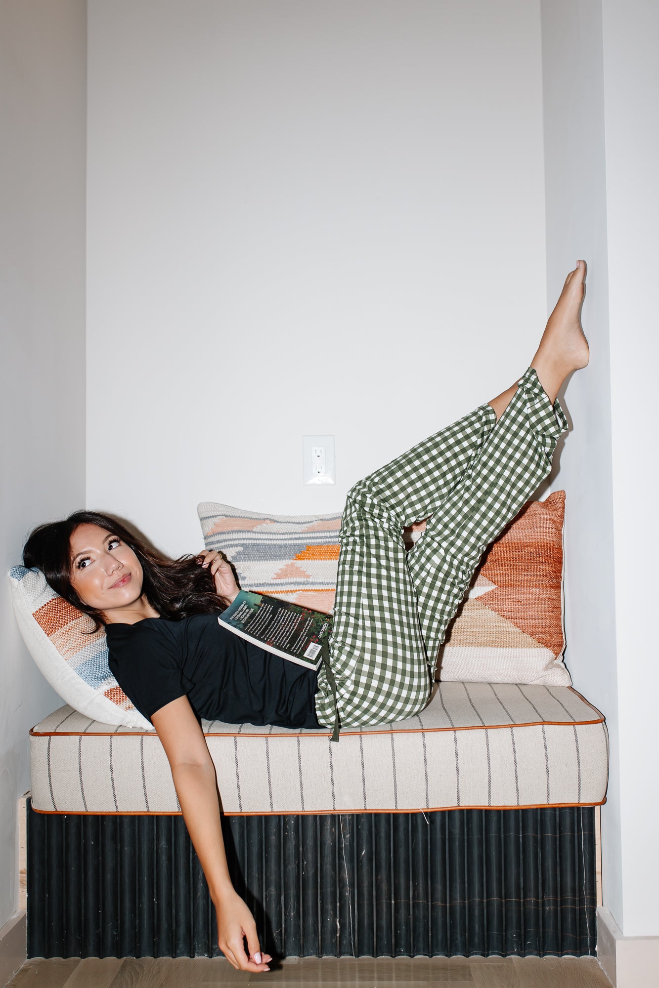 Model laying in a book nook while wearing the Women's Lounge Pants in Gingham Fir