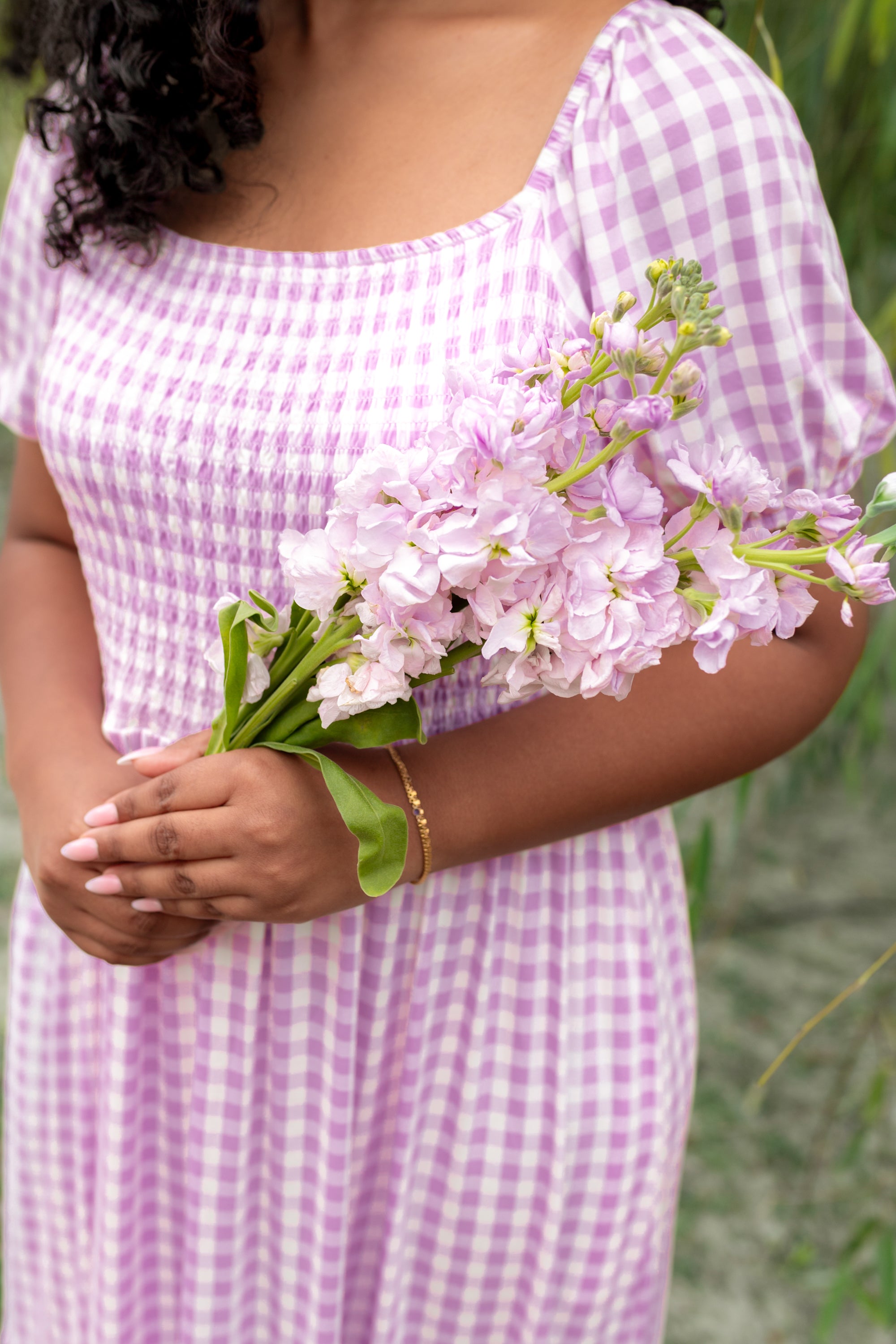 Person wearing a purple checkered dress holding pink flowers with a blurred green background
