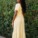 Woman wearing a yellow checkered dress standing against a green hedge.