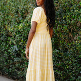 Woman wearing a yellow checkered dress standing against a green hedge.