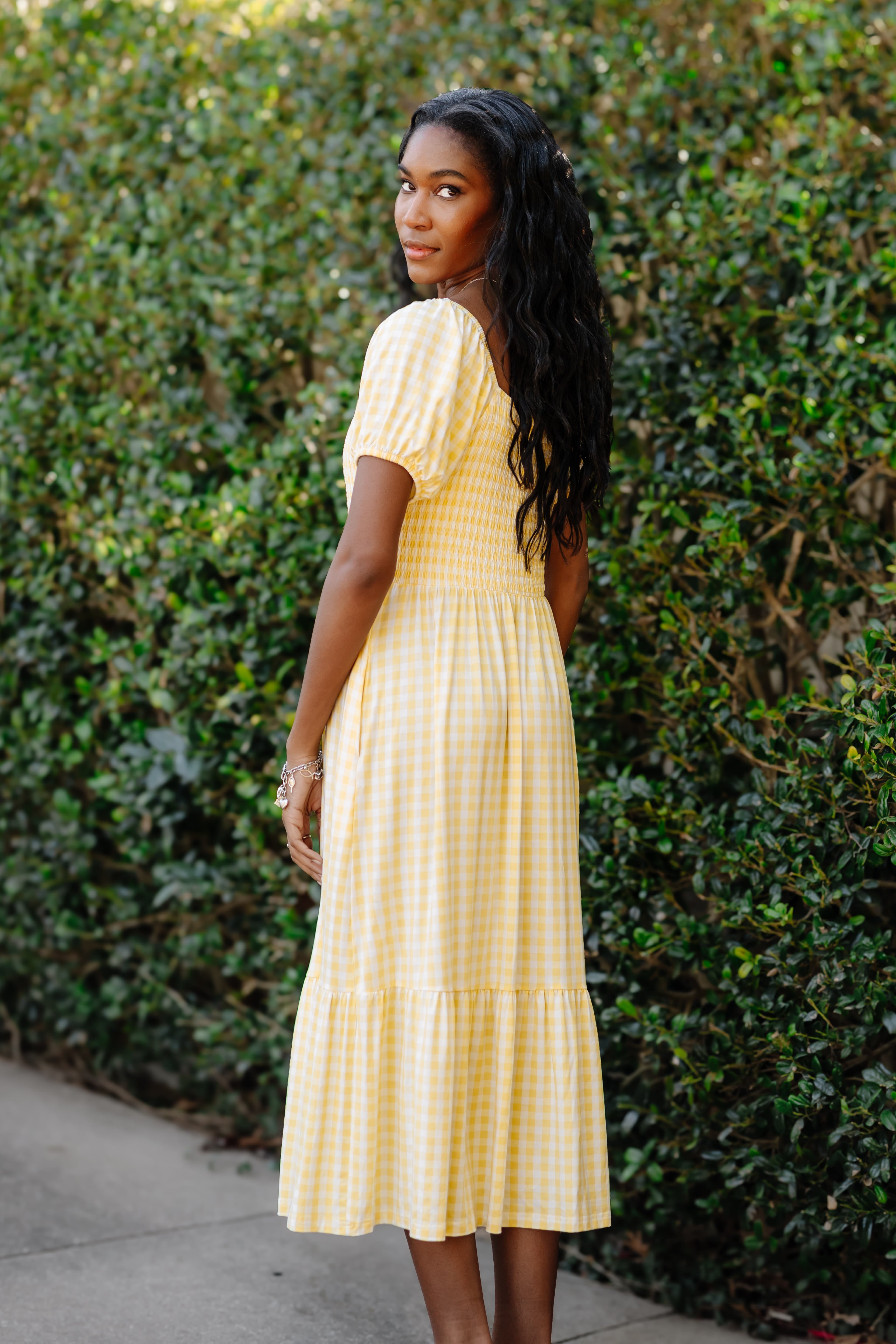 Woman wearing a yellow checkered dress standing against a green hedge.