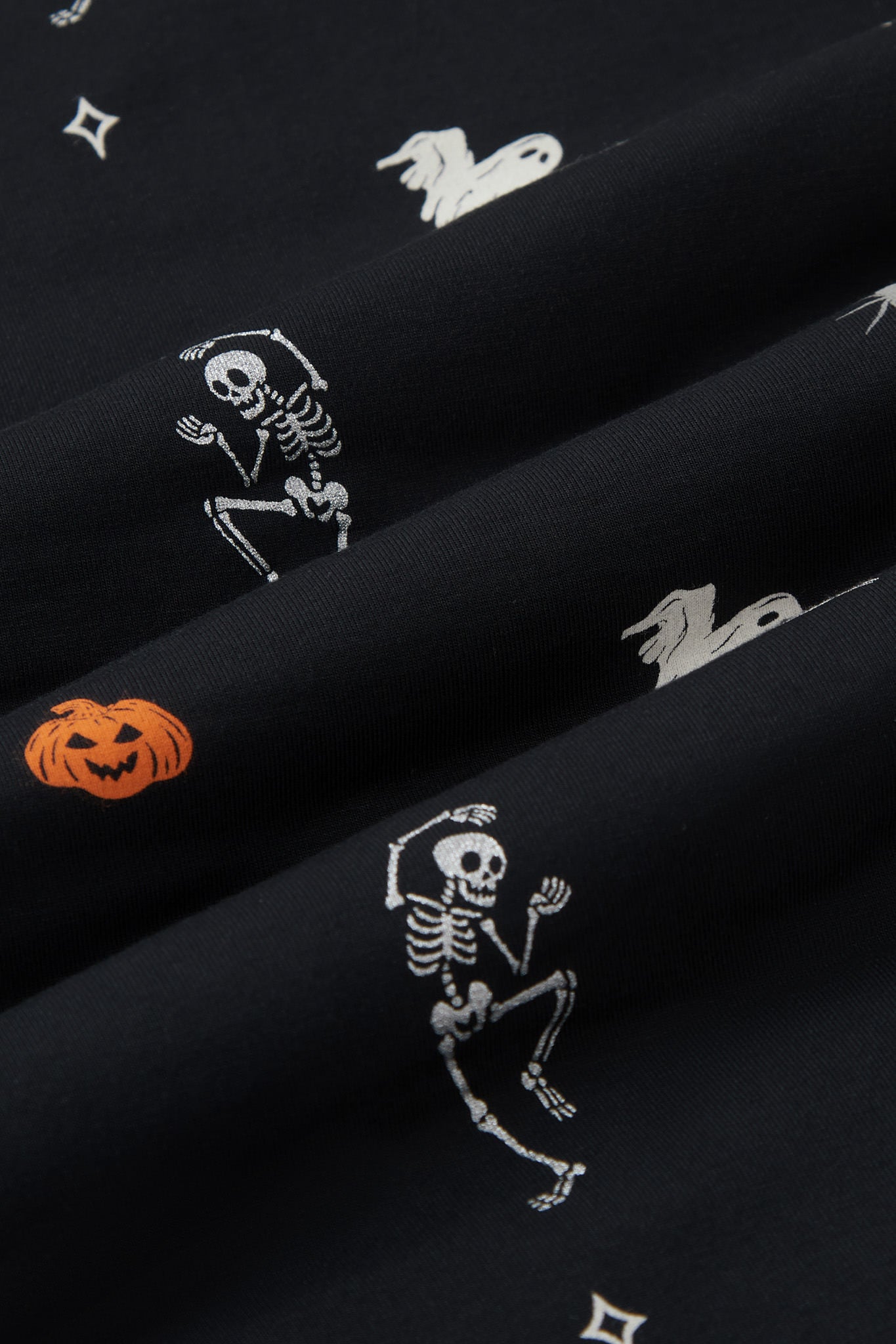 A repeating, all-over print featuring a flying white ghost, an orange jack o’ lantern, a dancing grey skeleton, a grey spider, and various white and grey twinkles, over a black background. ---1605 1