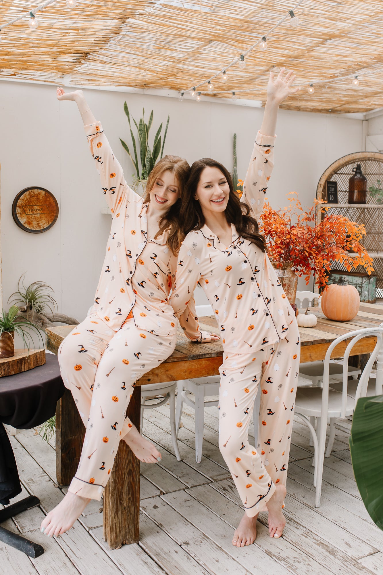 Two women in matching halloween pajamas sitting on a wooden table in a cozy room.