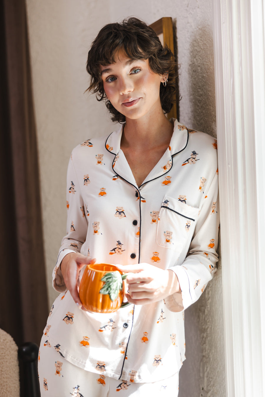 Modal standing against a wall holding a cup wearing a Beige Long-Sleeved Women's Pajama Set in Halloween Teddy print