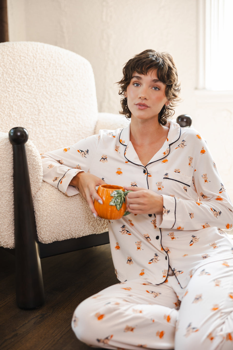 Modal sitting against a white sofa wearing Beige Long-Sleeved Women's Pajama Set in Halloween Teddy print while holding a mug
