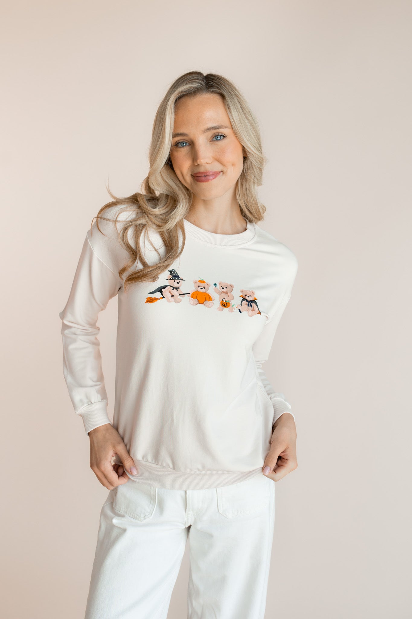 Modal posing in off white Women's Sweatshirt with Halloween Teddy print in the center over white pants standing in front of a beige background 