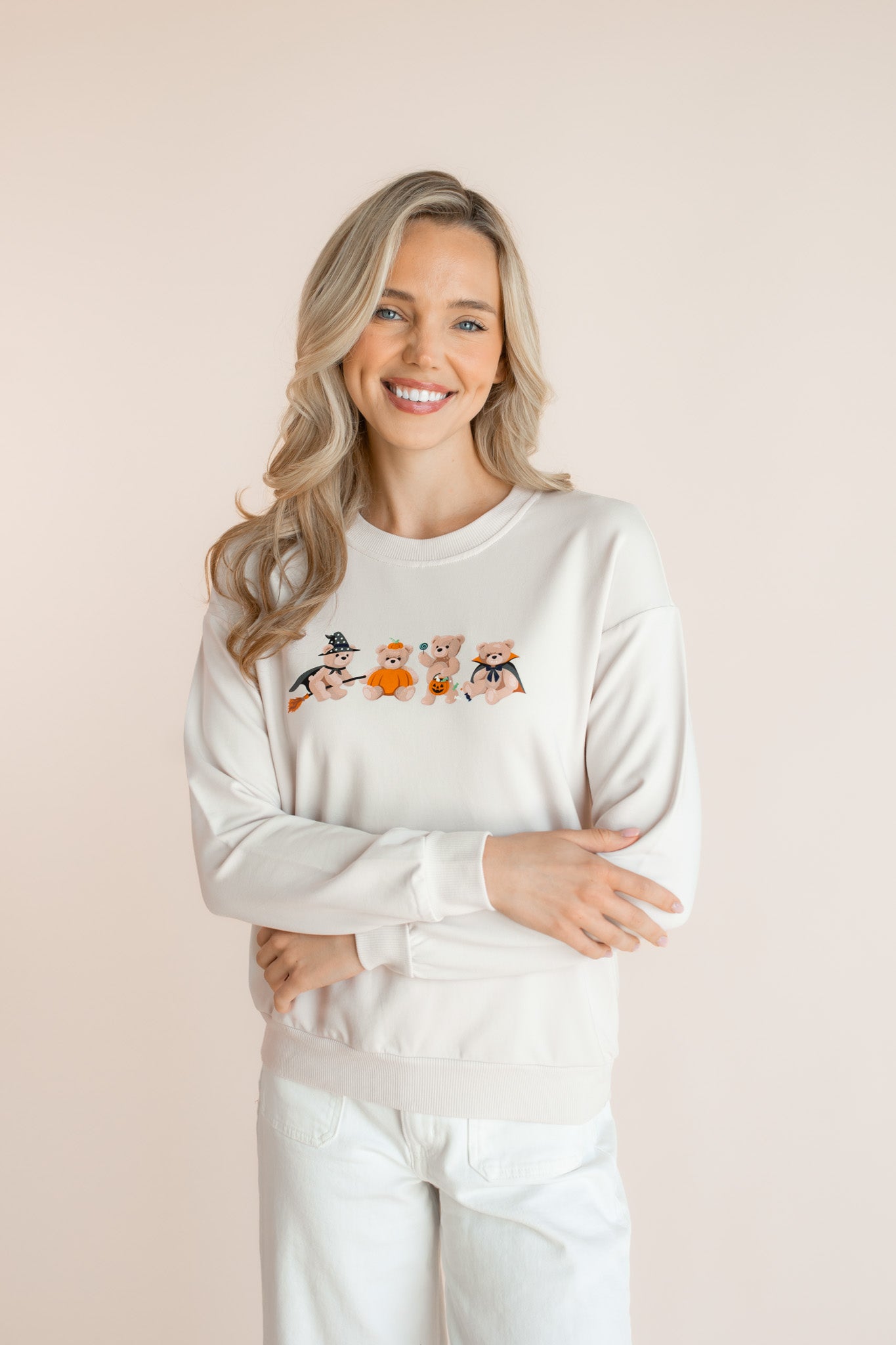 Modal wearing off white Women's Sweatshirt with Halloween Teddy print in the center over white pants smiling in front of a beige background