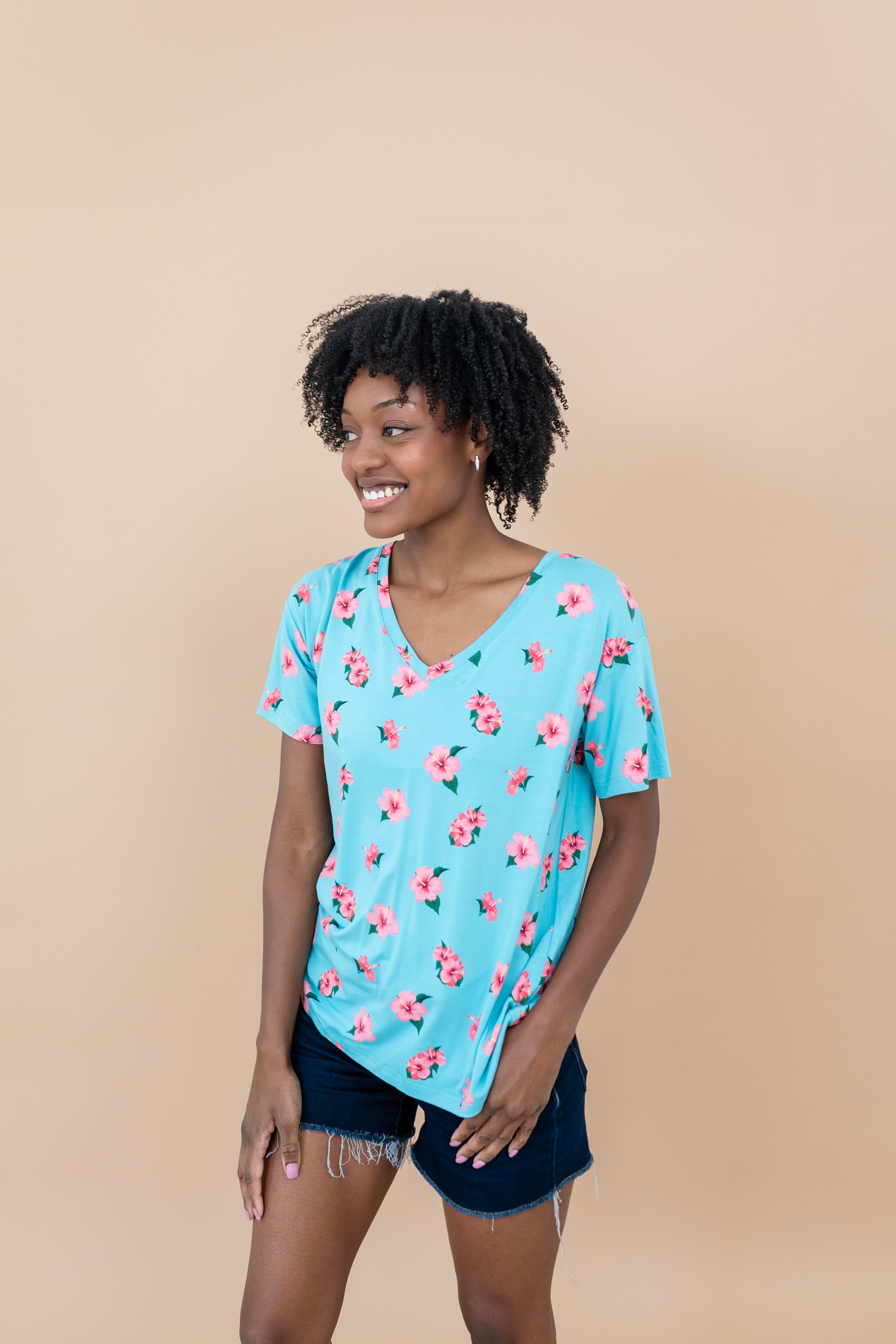 Woman wearing a blue floral Hibiscus v-neck tee and denim shorts against a beige background smiling 