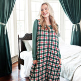 A woman stands in a bedroom wearing a green, red, and white plaid holiday sleep sack over a dark green long-sleeve top. The room features dark green curtains, a wooden bed, and hardwood floors.
::: Model is 5'9" wearing size XS ///