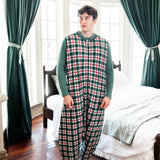 A man wearing a green, red, and white plaid sleep sack with solid green long sleeves, standing in a bedroom with dark green curtains and a bed in the background.