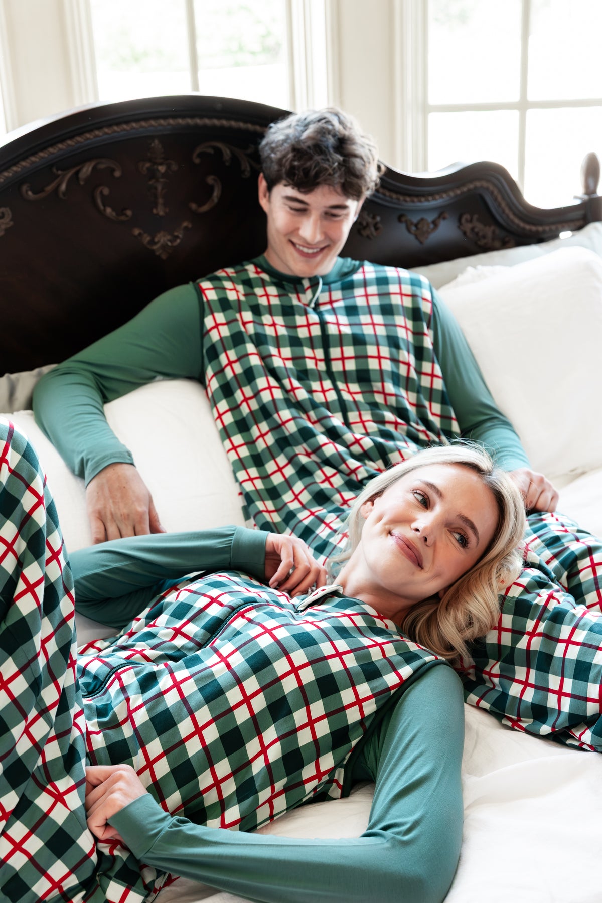 A man and a woman lying on a bed wearing matching green long-sleeve pajamas with red and white plaid sleep sacks