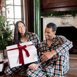 A man and woman wearing matching green, red, and white plaid pajamas sit on a wooden floor in a decorated living room, smiling while one holds a large white gift box with a red ribbon.