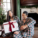 A man and woman wearing matching green, red, and white plaid pajamas sit on a wooden floor in a decorated living room, smiling while one holds a large white gift box with a red ribbon.