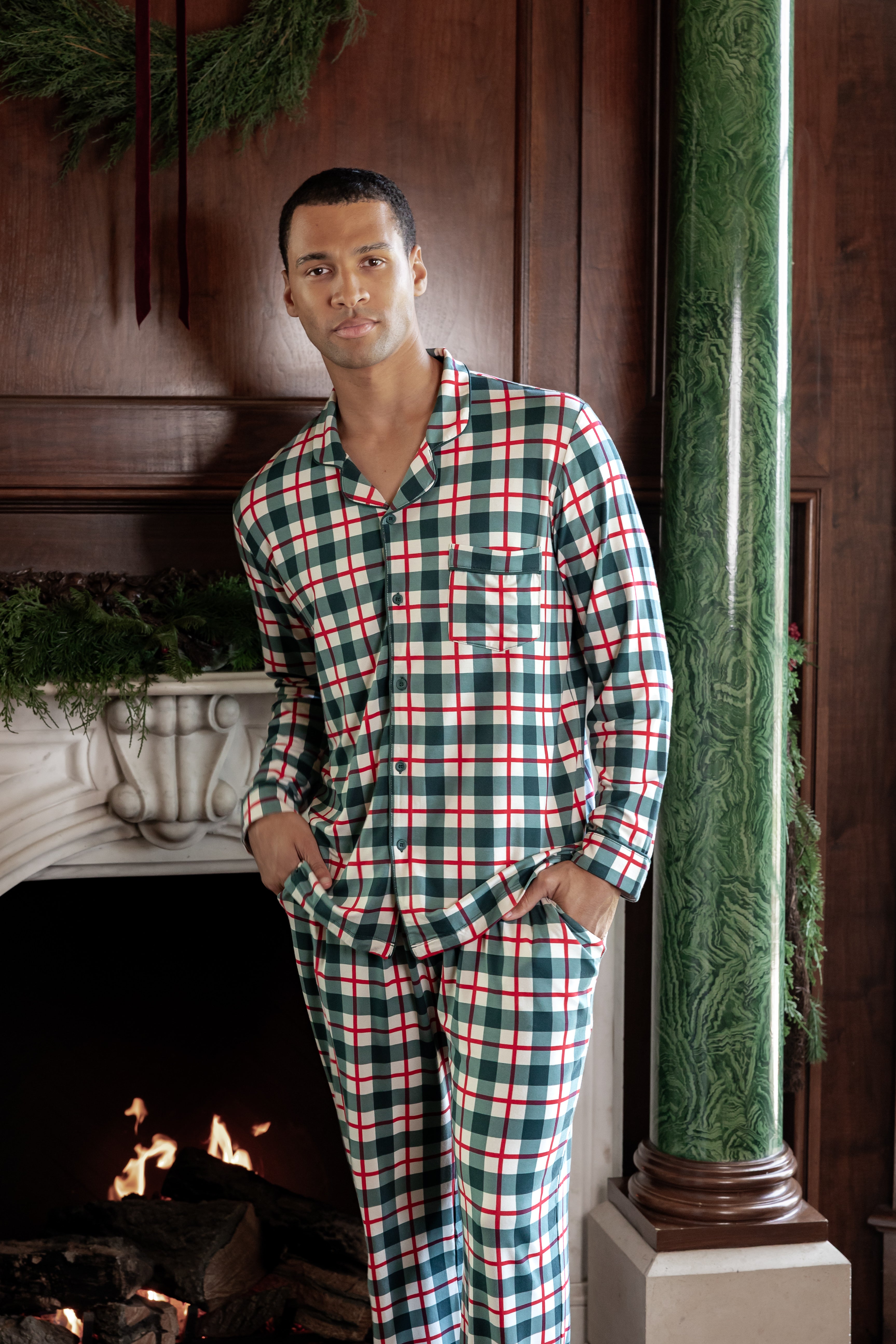 A man wearing a long-sleeve plaid pajama set in green, red, and white stands in front of a fireplace in a rustic room with dark wood paneling and a green marble column.
::: Model is 6'2" wearing size L ///
