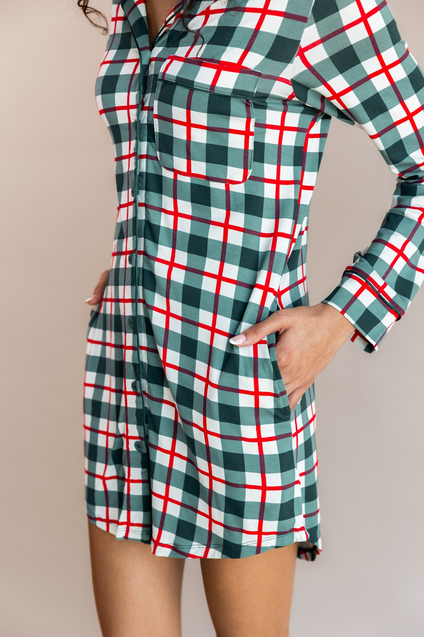 Woman standing to the side with her hands in her pocket wearing long sleeve sleep shirt in holiday plaid