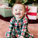 A smiling baby sitting on a patterned floor in front of a decorated Christmas tree and wrapped gifts, wearing a green, red, and white plaid long-sleeve pajama romper.