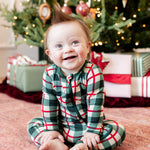 A baby sitting on a patterned rug in front of a decorated Christmas tree, wearing a green, red, and white plaid long-sleeve pajama set.