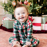 A baby sitting on a patterned rug in front of a decorated Christmas tree, wearing a green, red, and white plaid long-sleeve pajama set.