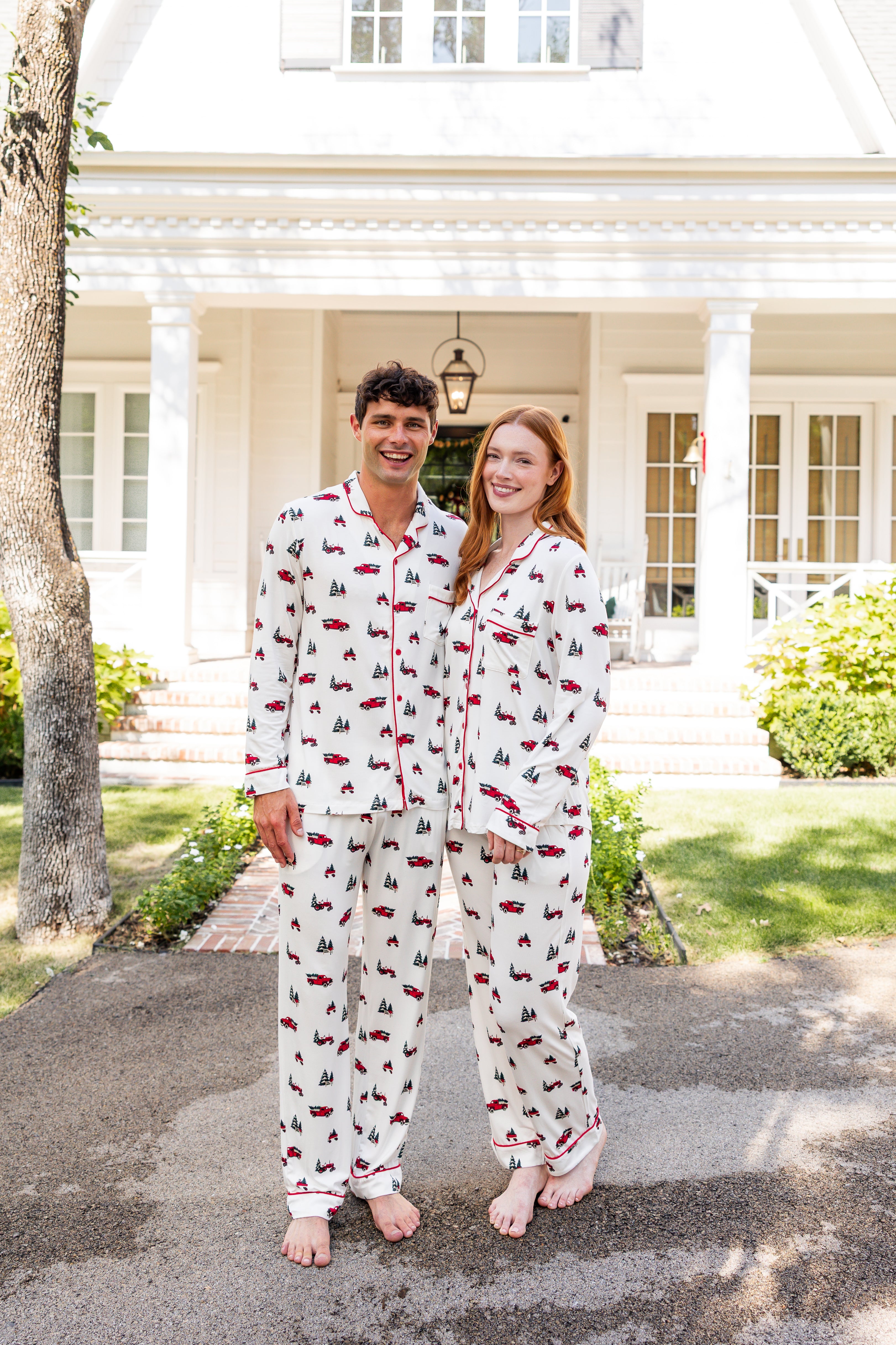Couple wearing matching christmas pajamas with red truck and christmas tree pattern in front of a white house