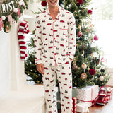A man wearing a white long-sleeve pajama set with a red and green Christmas tree and sleigh print, standing barefoot in a festive living room in front of a decorated Christmas tree.
::: Model is 6'1" wearing size M ///