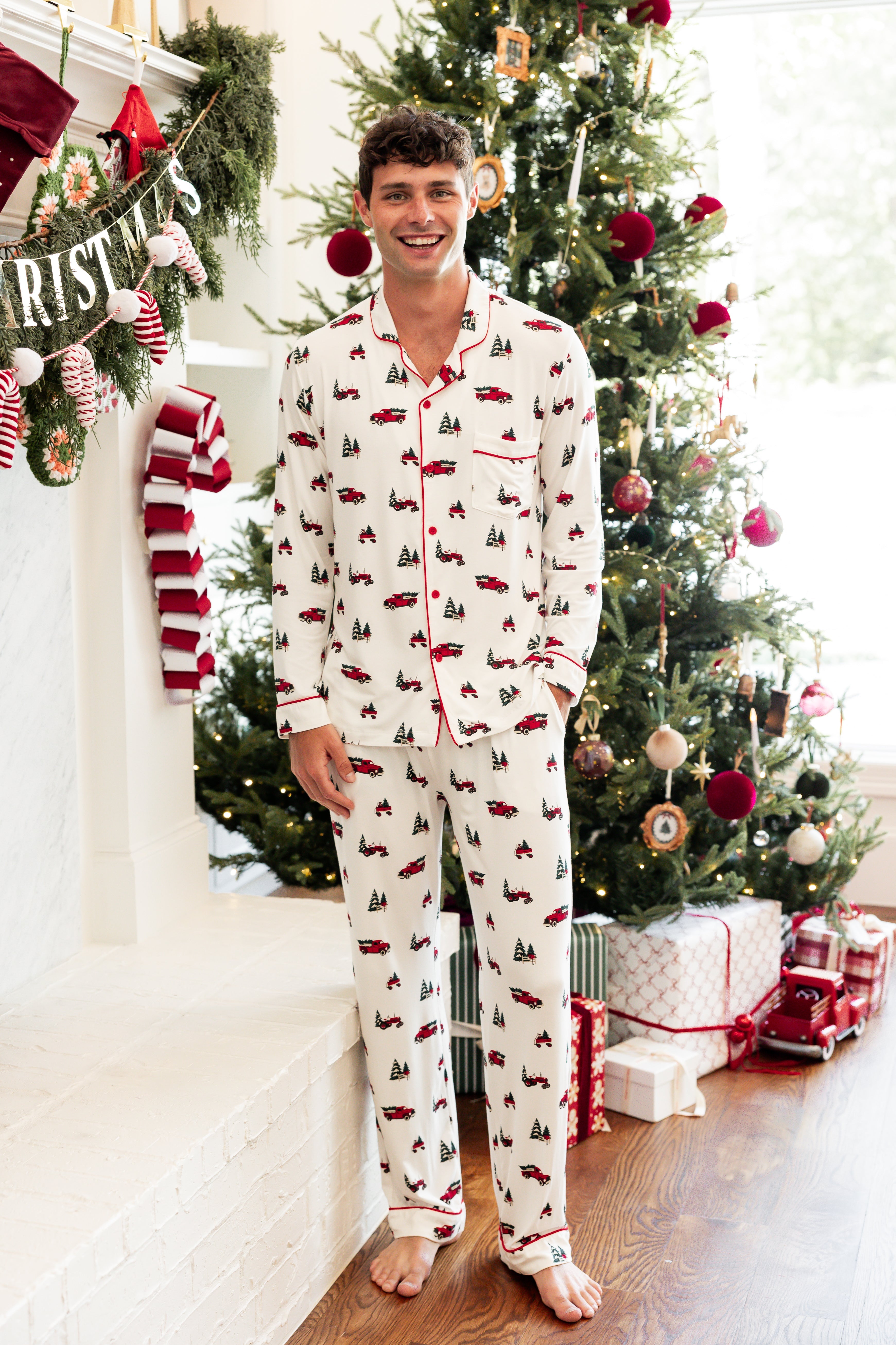 A man wearing a white long-sleeve pajama set with a red and green Christmas tree and sleigh print, standing barefoot in a festive living room in front of a decorated Christmas tree.
