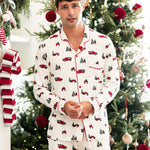 A man wearing a white long-sleeve pajama set with a red and green Christmas truck and tree print, standing in front of a decorated Christmas tree.
