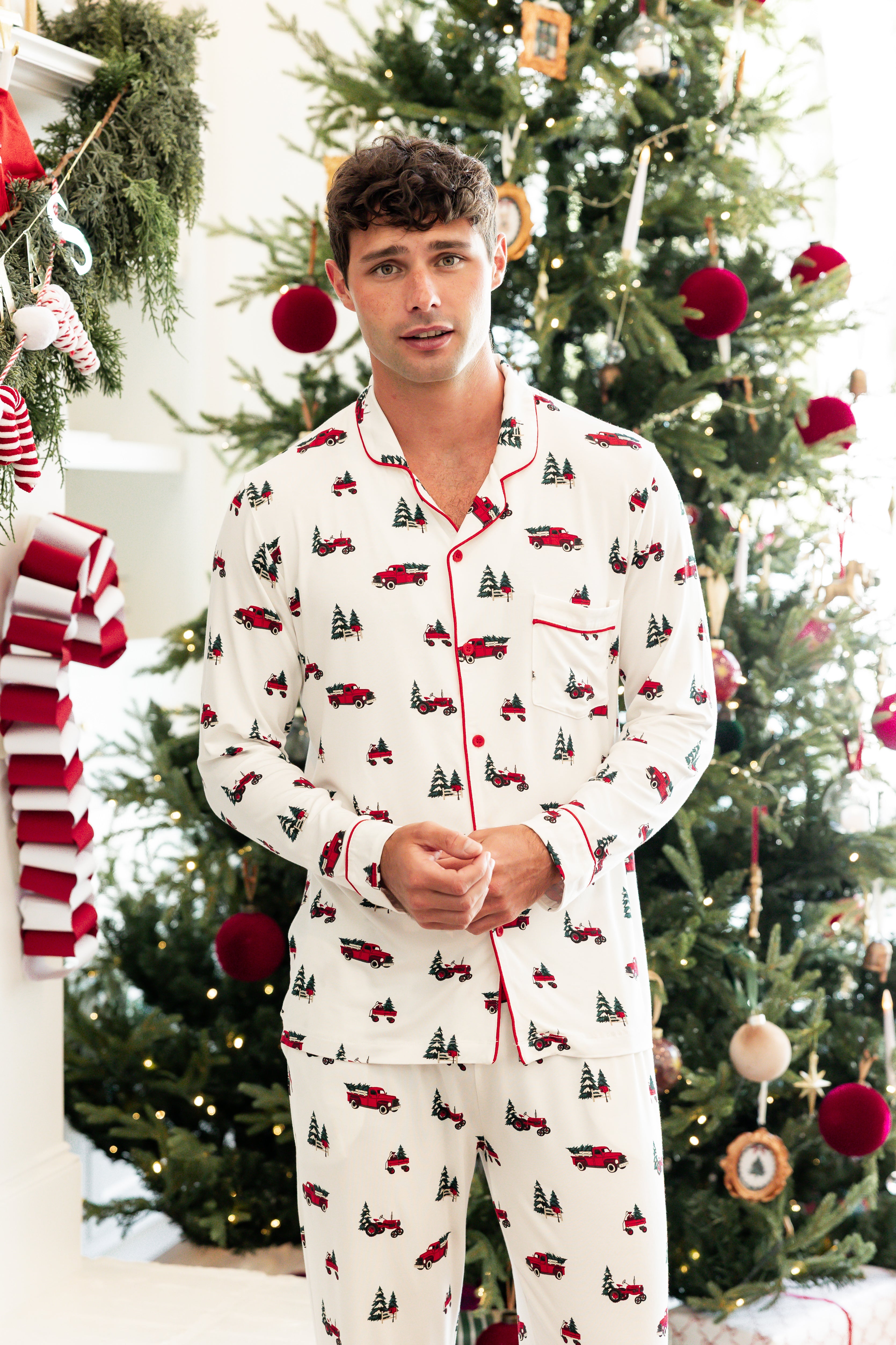 A man wearing a white long-sleeve pajama set with a red and green Christmas truck and tree print, standing in front of a decorated Christmas tree.
