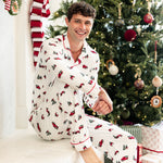 A man wearing a white Christmas-themed pajama set with red and green patterns sits on a white mantel in front of a decorated Christmas tree.