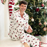 A man wearing a white Christmas-themed pajama set with red and green patterns sits on a white mantel in front of a decorated Christmas tree.