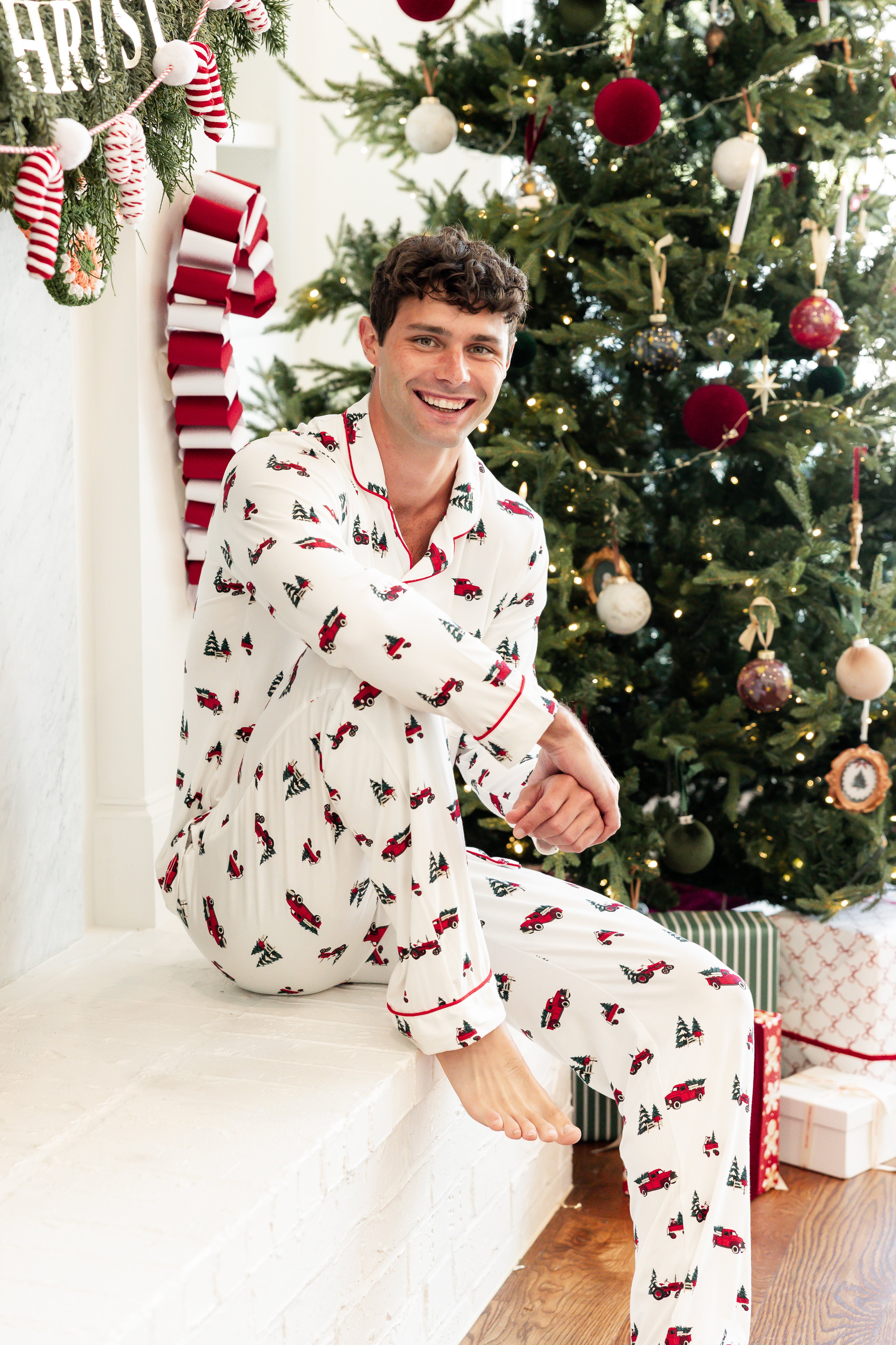 A man wearing a white Christmas-themed pajama set with red and green patterns sits on a white mantel in front of a decorated Christmas tree.