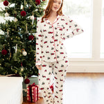 A woman wearing a matching holiday-themed pajama set stands barefoot in a living room next to a decorated Christmas tree. The pajamas are off-white with a repeating pattern of red trucks, green trees, and red accents.
---1605 3