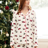 A woman with red hair wearing a cream-colored pajama set with a red and green Christmas truck and tree pattern, standing in front of a decorated Christmas tree.
::: Model is 5'8"" wearing size S ///
