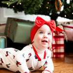 A baby wearing a white Christmas-themed romper with red trim and a matching red bow headband lies on a wooden floor under a decorated Christmas tree with wrapped presents.