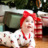 A baby wearing a white Christmas-themed romper with red trim and a matching red bow headband lies on a wooden floor under a decorated Christmas tree with wrapped presents.