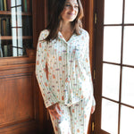 A woman wearing a long-sleeve button-up pajama set with a colorful Harry Potter Honeydukes pattern, standing in a sunlit room with wooden bookshelves and French doors.