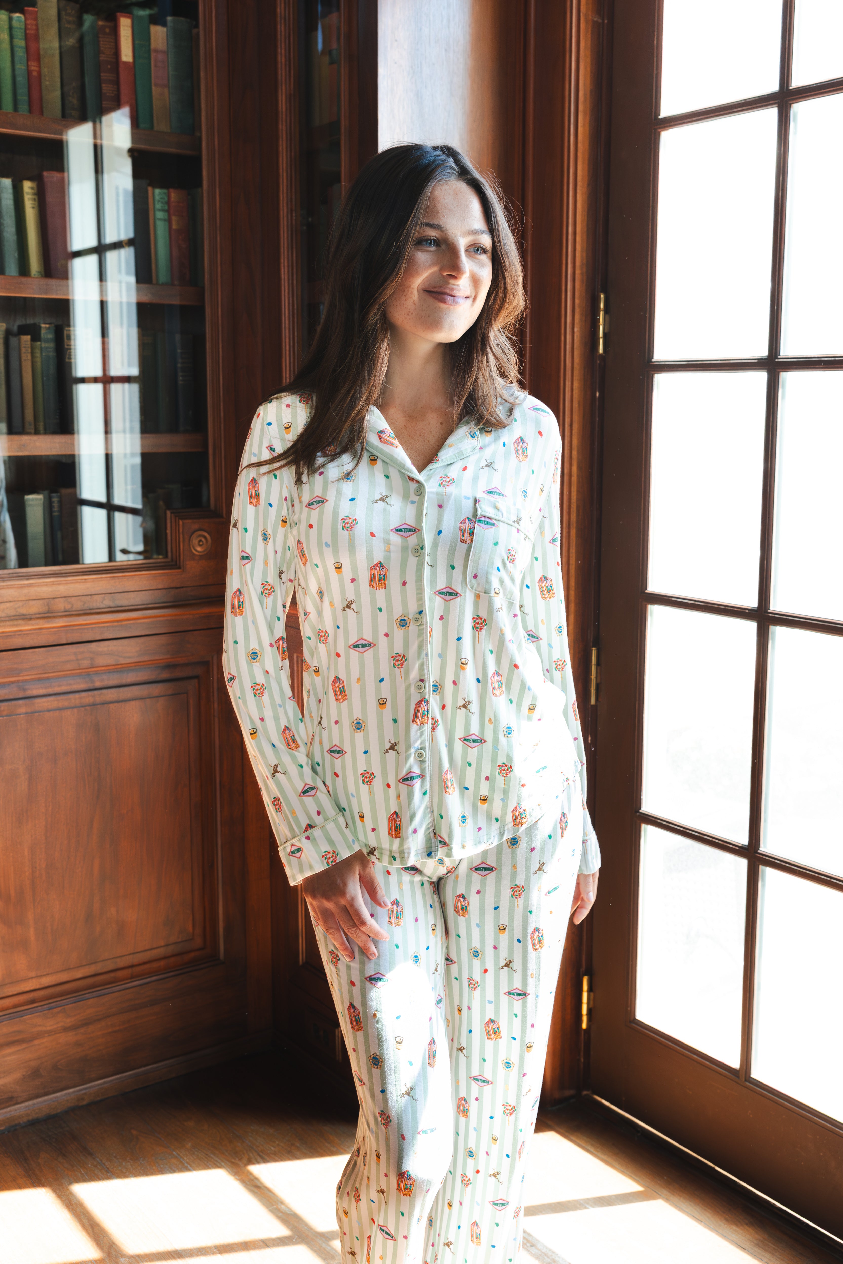 A woman wearing a long-sleeve button-up pajama set with a colorful Harry Potter Honeydukes pattern, standing in a sunlit room with wooden bookshelves and French doors.