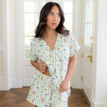 Woman in white room lifting bottom of top from the Women's Short Sleeve Pajama Set in Honeydukes and showcasing the waist band of the shorts   ::: Model is 5'2" wearing size S ///