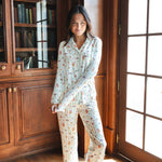 A woman stands barefoot in a sunlit room with wood bookshelves and glass doors, wearing a long-sleeve pajama set with a Harry Potter Honeydukes print on a light background.