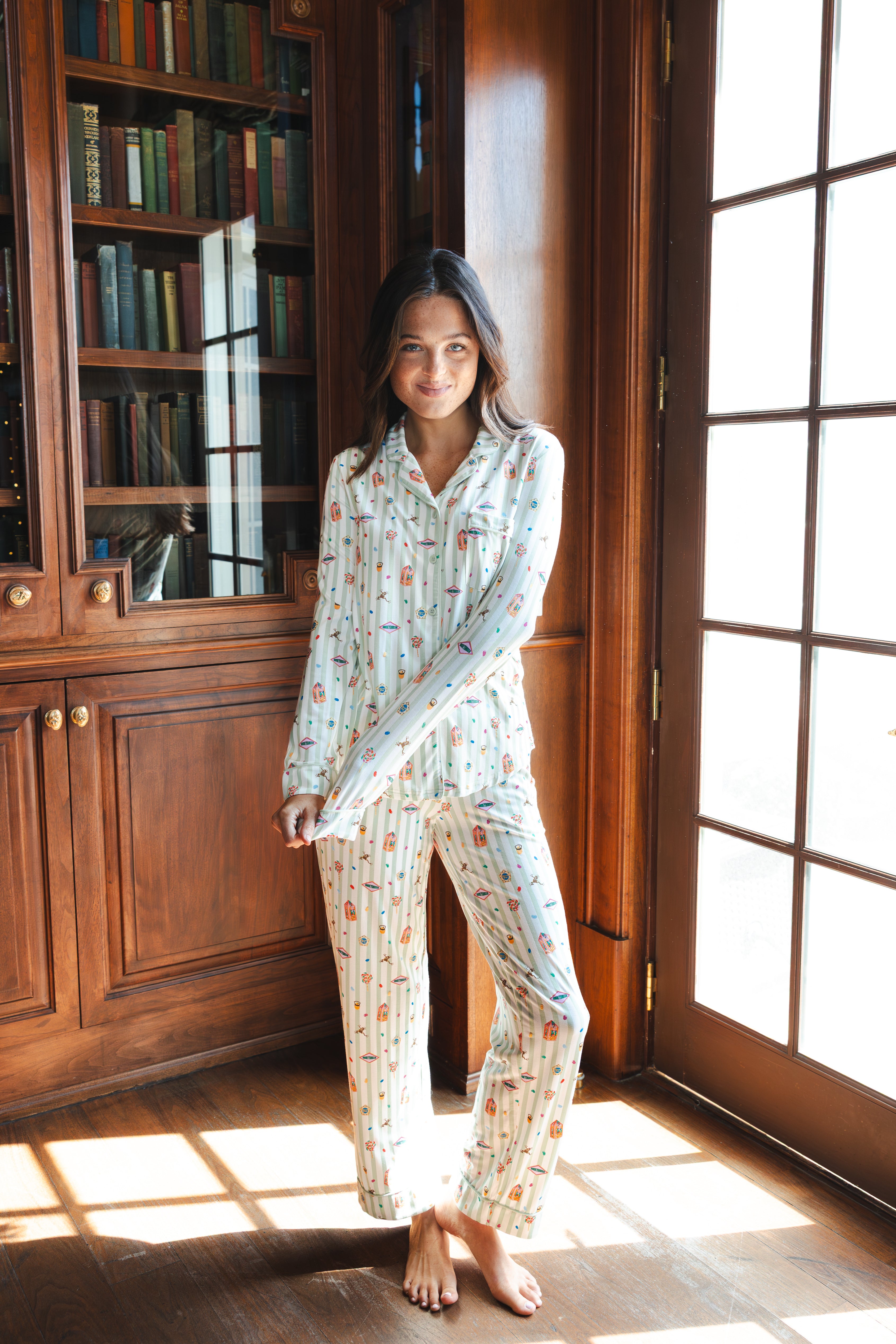 A woman stands barefoot in a sunlit room with wood bookshelves and glass doors, wearing a long-sleeve pajama set with a Harry Potter Honeydukes print on a light background.
