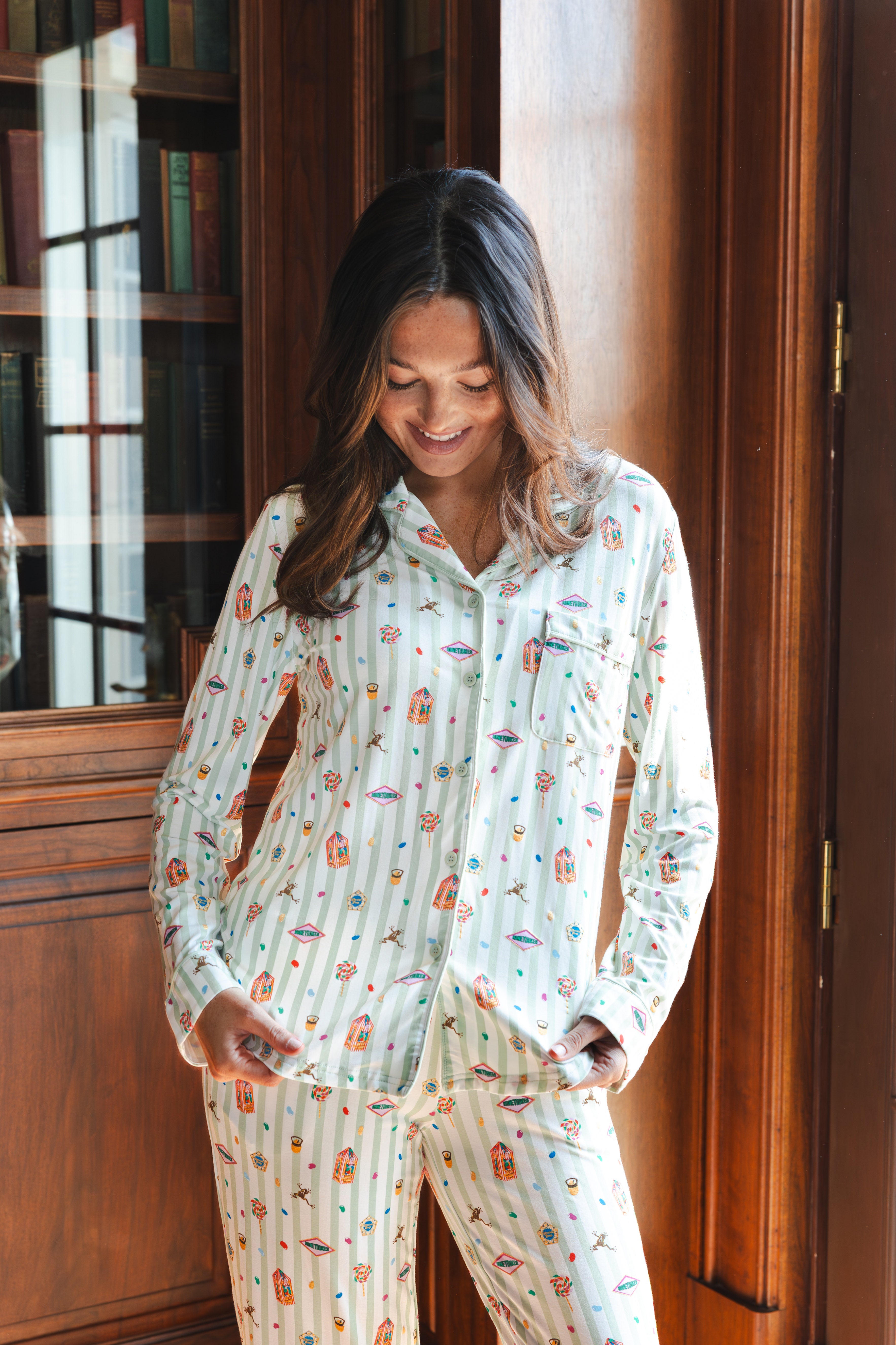 A woman wearing a long-sleeve pajama set with a colorful Harry Potter Honeydukes pattern on a white and light green striped background, standing in front of a wooden bookcase.