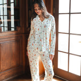 A woman wearing a long-sleeve button-up pajama set with a colorful Harry Potter Honeydukes pattern on a light background, standing barefoot in a sunlit room with wooden bookshelves and French doors.