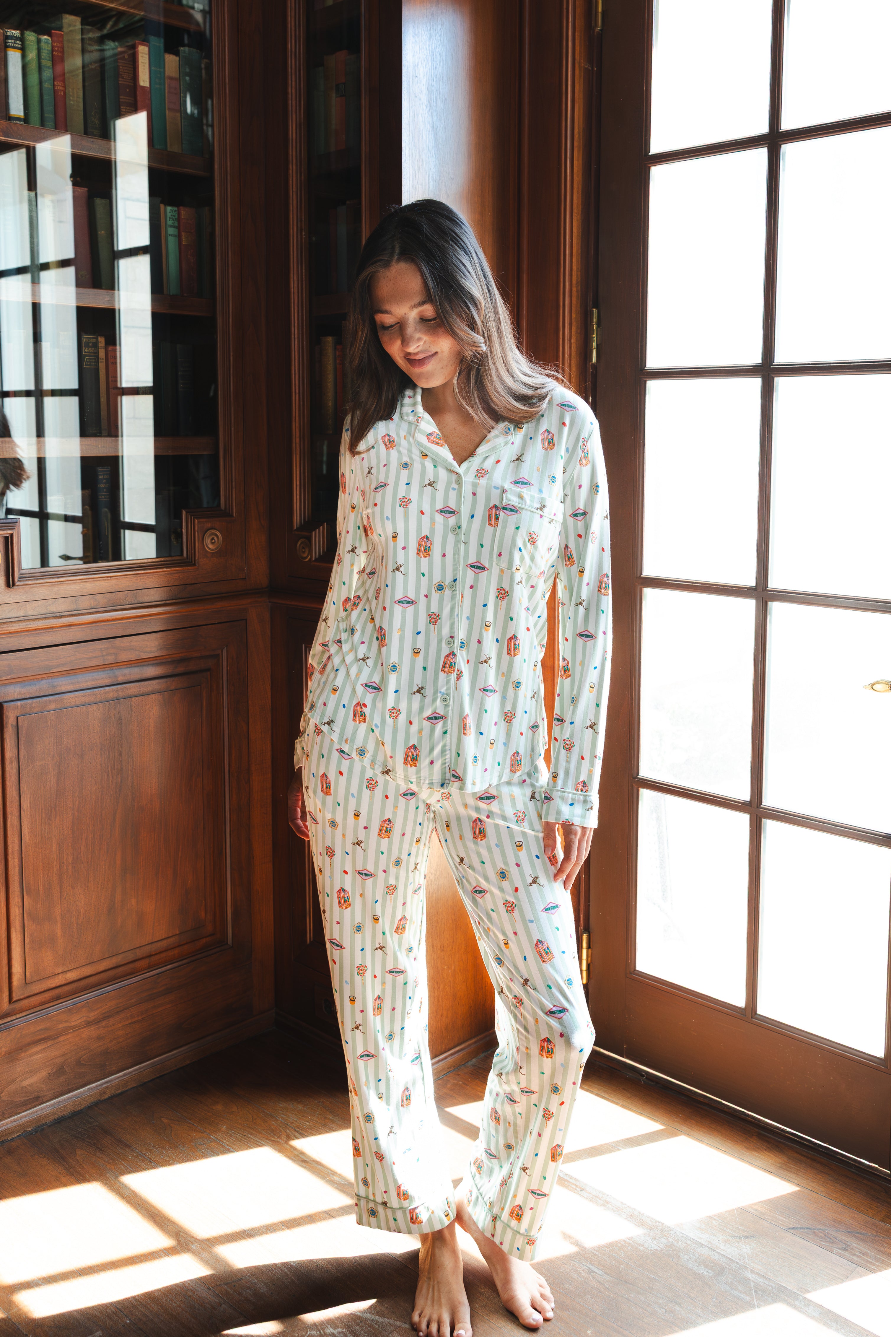 A woman wearing a long-sleeve button-up pajama set with a colorful Harry Potter Honeydukes pattern on a light background, standing barefoot in a sunlit room with wooden bookshelves and French doors.