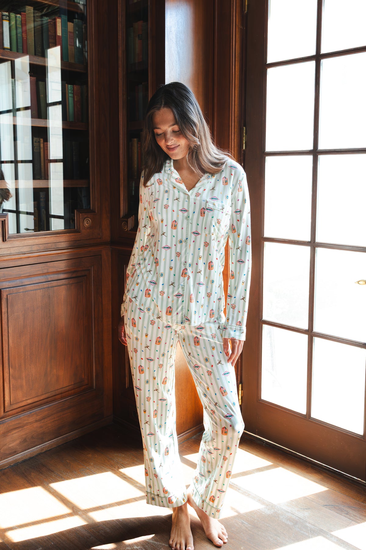 A woman wearing a long-sleeve button-up pajama set with a colorful Harry Potter Honeydukes pattern on a light background, standing barefoot in a sunlit room with wooden bookshelves and French doors.