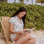 Model sitting on a rattan beach chair while reading and wearing the Kyte Short Sleeve Pajama Set in Dew Surf