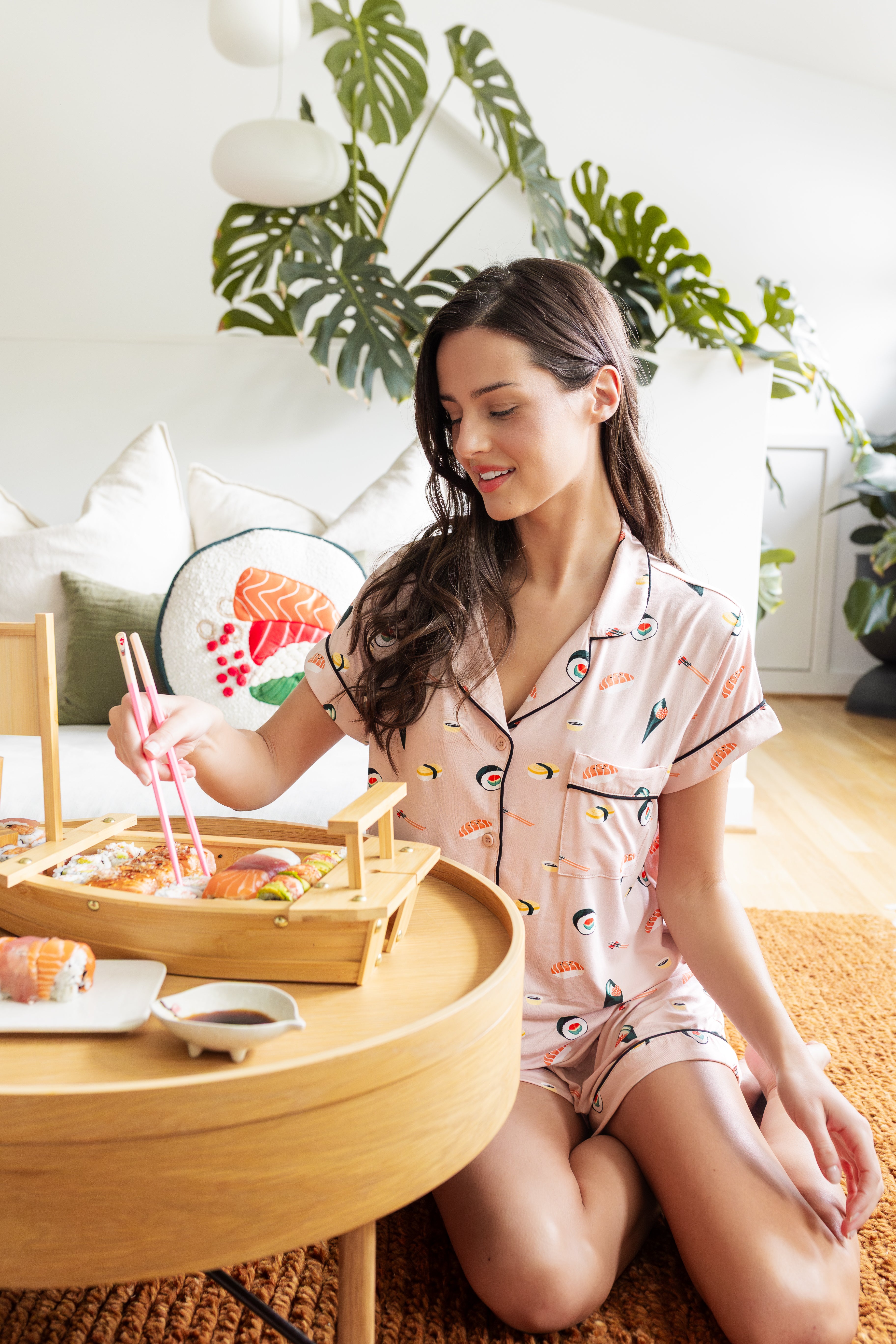 Woman in pink sushi themed pajamas sitting on the floor with a tray of food in a cozy room.
---PJ 2