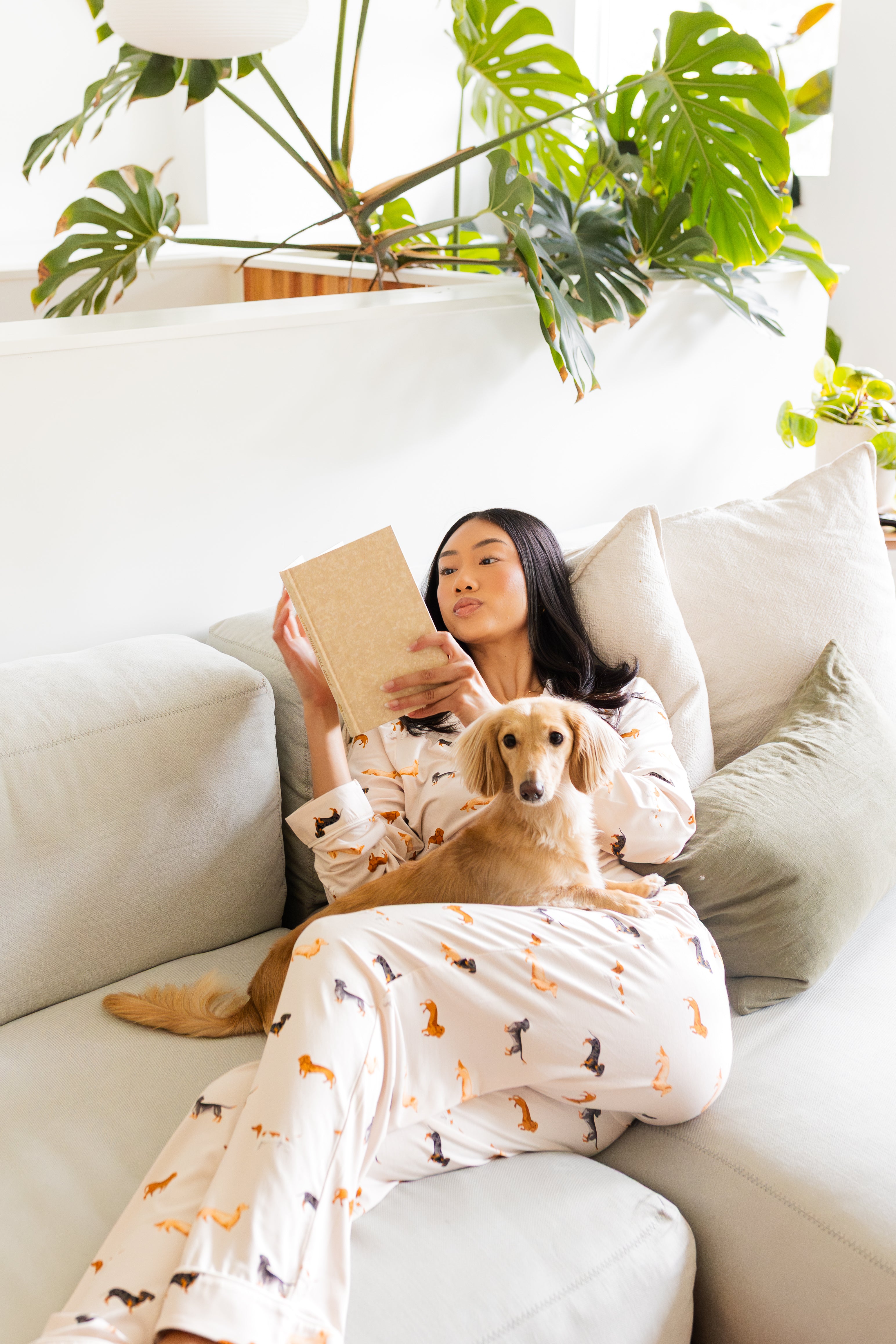 Woman wearing Long-Sleeved Women's Pajama Set in Dachshund reading a book to her blonde dachshund  on a couch with plants in the background

---1605 3