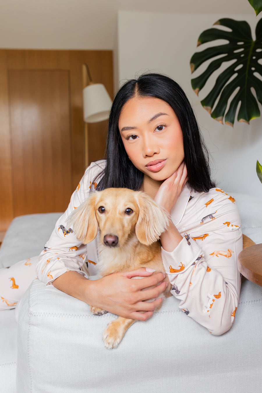 Woman in Long-Sleeved Women's Pajama Set in Dachshund holding a blonde dachshund on a couch with a plant in the background