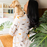 Back side of model wearing the Long-Sleeved Women's Pajama Set in Dachshund while holding a blonde dachshund with a large green plant 